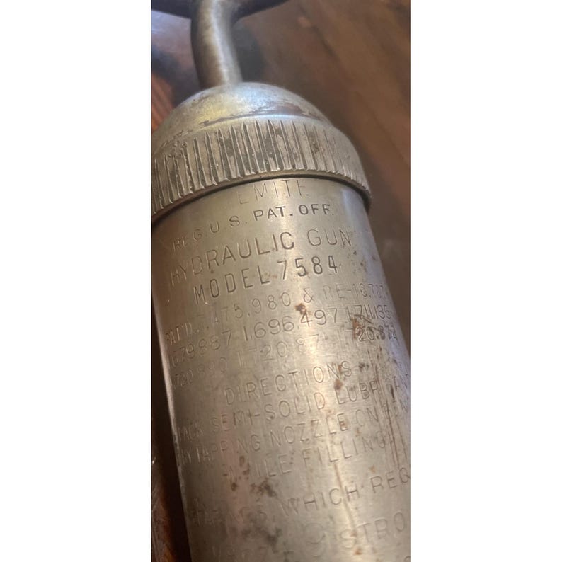 Pode incluir: Uma pistola hidr&aacute;ulica vintage, modelo 7584, com texto vis&iacute;vel, incluindo "REG. U.S. PAT. OFF." e "MITE." A pistola de metal tem um acabamento prateado e um bico curvo. O corpo da pistola exibe instru&ccedil;&otilde;es e informa&ccedil;&otilde;es de patentes.