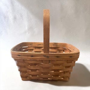 May include: A rectangular woven wood basket featuring a light brown finish and a single upright handle. The basket is constructed with flat, interlaced wooden splints and is secured with small dark metal nails along the top rim and handle base.