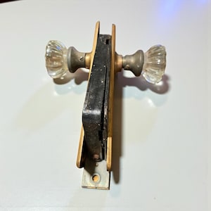 May include: An antique door lock with two clear glass doorknobs. The lock mechanism is black and gold, with a metal plate for mounting. The doorknobs have a ribbed design and are attached to the lock with brass hardware.
