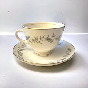 May include: Cream-colored teacup and saucer set with a floral design. The cup and saucer have a gold trim. The floral pattern includes small pink and white flowers with green leaves. The cup has a handle and sits on a small pedestal.