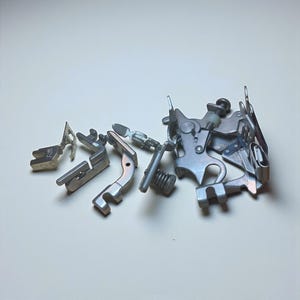 May include: A collection of silver-colored sewing machine presser feet and related parts, scattered on a white surface. The parts include various shapes and sizes, with intricate mechanical details.