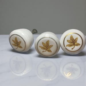 May include: Three white ceramic cabinet knobs with a gold leaf design. Each knob has a gold rim and a brass stem. The knobs are arranged on a reflective surface.