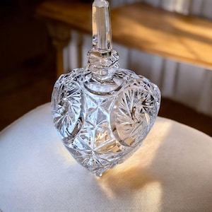 May include: A clear, heart-shaped crystal jar with a faceted design and a tall, pointed stopper. The jar has intricate patterns and is likely used for decorative storage.