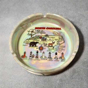 May include: A vintage ashtray with a map of North Carolina, featuring landmarks like lighthouses and a bear. The ashtray has a pearlescent finish and a circular shape. The text "North Carolina" is displayed prominently.