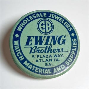 May include: A round, vintage metal container with a light green background and blue text. The text reads "WHOLESALE JEWELERS" around the edge, with "EWING Brothers" in the center. The container also says "WATCH MATERIAL AND SUPPLIES" and "5 PLAZA WAY, ATLANTA, GA."