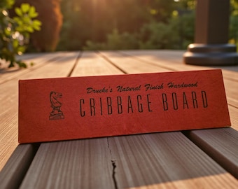May include: A red cribbage board with the text "Drueke's Natural Finish Hardwood CRIBBAGE BOARD" and a knight chess piece logo. The board is on a wooden surface, with sunlight in the background.