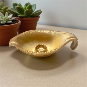 May include: A gold-colored ceramic candle holder with the text "CHRIST THE LIGHT OF THE WORLD" inscribed around the rim. The holder has a curved handle and a central well for a candle. Succulent plants in terracotta pots are in the background.