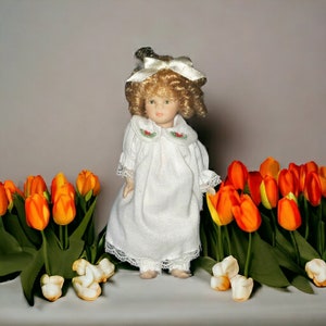 May include: A porcelain doll with blonde curly hair and a white dress with lace trim stands among orange and yellow tulips. The doll has a white bow in her hair and a painted face. The tulips have green leaves.