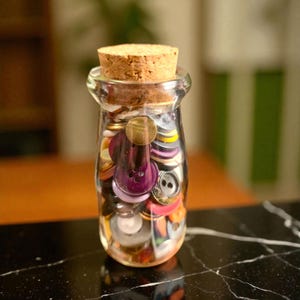 May include: A clear glass jar filled with a variety of colorful buttons, sealed with a cork stopper. The buttons are in various sizes, shapes, and colors, including purple, gold, and silver. The jar sits on a black and white marble surface.