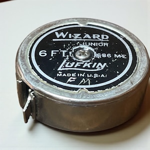 May include: A vintage Lufkin Wizard Junior tape measure. The tape measure is 6 feet long. The face of the tape measure is black with white lettering. The tape measure is made in the U.S.A.