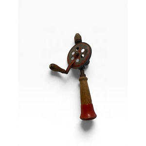 May include: A vintage hand drill featuring a red metal gear with circular cutouts, a wooden side handle, and a vertical wooden grip with a red base. The tool includes a metal chuck at the top for holding drill bits, showing signs of wear.