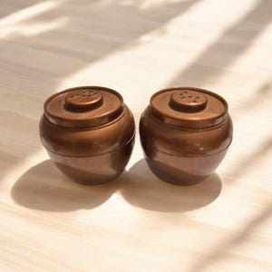May include: Two brown, round containers with lids. The containers have a smooth, glossy finish and are sitting on a light-colored wooden surface. The lids have a small, circular design on top. The containers are casting shadows.