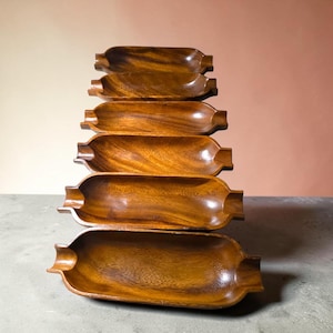 May include: A stack of six elongated, oval-shaped wooden trays with a smooth, polished finish. Each tray features a deep, concave center and small, notched indentations on both ends, showcasing a rich, natural brown wood grain pattern throughout.