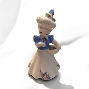 May include: A vintage ceramic figurine of a woman in a dress and hat, holding a basket. The figurine is painted in pastel colors, with floral accents on the dress. The woman's hat is decorated with flowers.