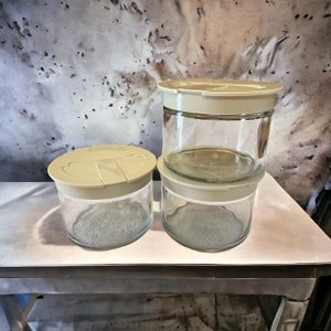 May include: Three clear glass jars with beige lids are stacked on a white surface. The jars are cylindrical and appear to be empty. The lids have a simple, functional design. The background is a mottled pattern of gray and white.