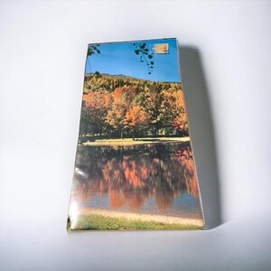 May include: A rectangular box with a scenic image of a lake reflecting autumn trees under a blue sky. The trees display vibrant orange, red, and yellow foliage. A small sticker is visible on the top right corner.