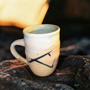 May include: A ceramic mug with a two-tone finish, featuring a creamy off-white upper section and a warm tan lower section. The exterior is decorated with a bold, black hand-painted angular line design. The mug has a curved handle and a smooth rim.