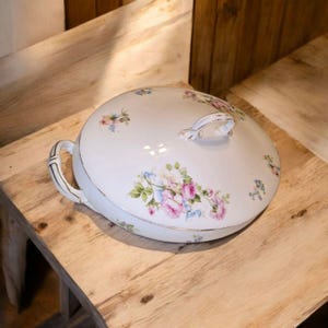 May include: A white ceramic serving dish with a lid, adorned with pink, blue, and green floral patterns. The dish has a handle on the lid and a handle on the side. The rim is trimmed with gold.