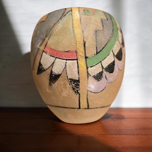 May include: A rounded, tan-colored ceramic vessel featuring a hand-painted design with a central vertical yellow stripe. The surface displays geometric patterns in shades of gray, red, green, and black, with a series of petal-like shapes along the sides.