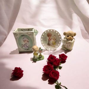 May include: A collection of Precious Moments figurines and a small plate with a floral design. The figurines include a bear, a girl holding a sign, and a figure inside a decorative holder. Red roses are arranged in the foreground.