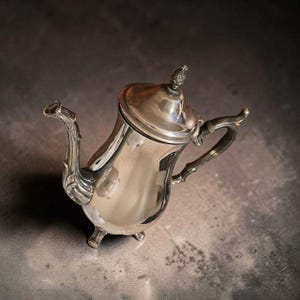May include: A polished silver-toned teapot featuring a curved spout, a handle with ornate detailing, and a lid topped with a small, textured finial. The teapot stands on three small, curved feet and has a smooth, reflective metallic surface.