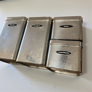 May include: A set of four rectangular, stainless steel canisters for kitchen storage. Each canister has a black oval label with white text: "flour", "sugar", "tea", and "coffee". The canisters are arranged in a row.