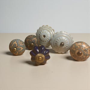 May include: A collection of decorative ceramic cabinet knobs in various colors and designs. The knobs include floral and patterned details, with colors like blue, gold, and purple. Each knob has a metal screw for attachment.