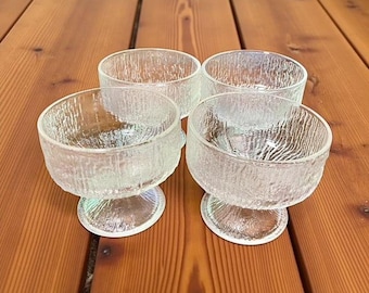 May include: Four clear glass dessert bowls with textured exteriors and pedestal bases are arranged on a wooden surface. The bowls have a round shape and are designed for serving desserts or small portions.
