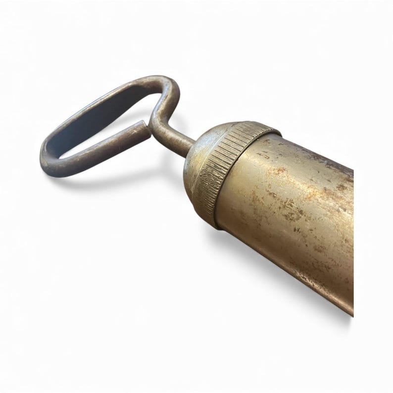 Pode incluir: Close-up de uma pistola de graxa antiga com uma al&ccedil;a curva e um corpo cil&iacute;ndrico. O metal tem uma apar&ecirc;ncia manchada e envelhecida, com desgaste vis&iacute;vel. A al&ccedil;a tem formato de la&ccedil;o.