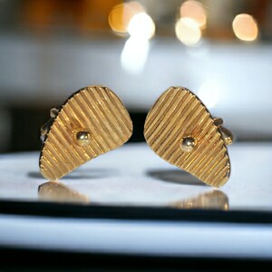May include: A pair of gold-colored cufflinks. Each cufflink has a unique shape with a textured, ridged surface and a small, round, gold-colored accent. The cufflinks are displayed on a reflective surface.