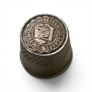 May include: A silver thimble with the text "M. Mitchell Elevation 6684 ft. N.C." encircling a central emblem. The thimble has a textured surface and a decorative rim.