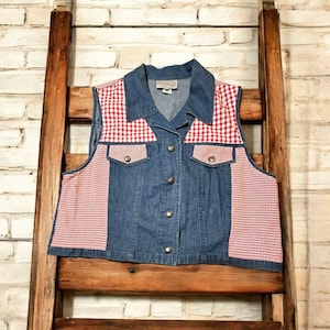 May include: A denim vest with red and white gingham accents. The vest features a collared neckline, two front pockets, and gold-colored buttons. The red and white gingham fabric is used on the shoulders and side panels.