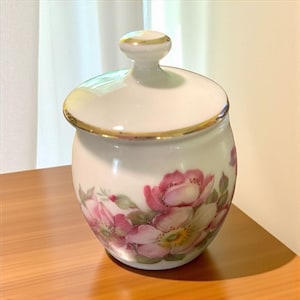 May include: A white ceramic jar with a lid, adorned with pink floral designs and gold trim. The jar has a rounded shape and a small knob on the lid. The floral pattern features pink roses and green leaves.