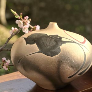 May include: A rounded ceramic vase with a matte beige finish, featuring a prominent black abstract floral design and a thin red curved line. The vessel has a narrow opening at the top and a textured surface, showcasing a hand-crafted appearance.