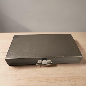 May include: A rectangular, gray metal box with a hinged lid and a silver-colored clasp and handle. The box is sitting on a light-colored wooden surface.