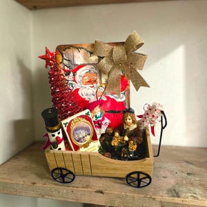 May include: A wooden wagon filled with Christmas decor including a red glittery tree, a Santa Claus illustration, a small nutcracker, a book titled Christmas Stories, a figurine of a person, and a large burlap bow attached to the back.