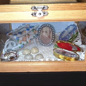 May include: A wooden display box with a glass front, showcasing various decorative items. Inside are a porcelain wheelbarrow, a framed portrait, seashells, and a small, gold-trimmed box with a red interior. The box is secured with a silver clasp.