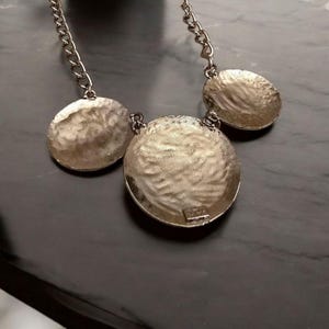 May include: A silver-toned necklace featuring three textured, circular pendants. The pendants are connected by a chain. The necklace has a hammered metal appearance, reflecting light. The design is simple yet elegant, suitable for various occasions.