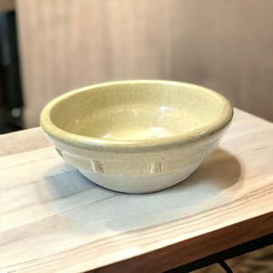 May include: A light yellow ceramic bowl with a slightly textured surface. The bowl has a rounded shape with a wide rim and a series of rectangular indentations around the lower portion. The bowl is sitting on a light-colored wooden surface.