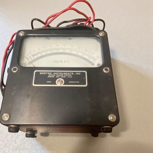 May include: A vintage black Weston Instruments voltmeter with a white gauge face displaying voltage readings. The meter has a needle indicator and the text "VOLTS A.C." is visible. Red and black wires extend from the top. The text "WESTON INSTRUMENTS, INC." is printed on a label.