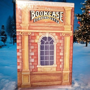 May include: A sealed deck of playing cards with a decorative design. The box features a brick facade with a window and columns, and the text "BOOKCASE PLAYING CARDS" in a banner above the window.