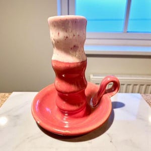May include: A handmade ceramic candle holder in shades of red and white. The candle holder has a round base with a handle and a tall, cylindrical cup to hold a candle. The top of the cup is white with red speckles.