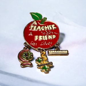 May include: A red apple-shaped pin with the text "A Teacher Today... A Friend for Life!" and dangling charms of a globe, math symbols, and a ruler. The pin has gold-colored trim and green accents.