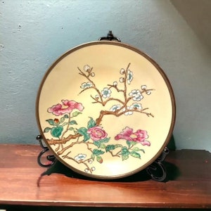 May include: A round decorative plate with a floral design. The plate features pink flowers and white blossoms on a beige background, with a brown border. It is displayed on a black metal stand, set against a blue wall and a wooden surface.