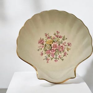May include: A cream-colored, shell-shaped decorative plate with a gold rim. The plate features a floral bouquet in the center with pink, yellow, and green flowers. The plate is sitting on a white surface.