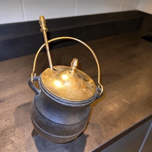 May include: A black cast iron pot with a rounded body and three small feet. It features a polished brass lid with a central knob, a matching brass swing handle, and a long, slender brass spoon resting inside through a small notch in the lid.