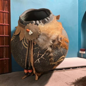 May include: A round, dark brown ceramic vessel with a wide opening, featuring a textured surface with light-colored line patterns. It is decorated with a tan leather star-shaped patch, orange and white accents, brown feathers, and leather fringe.