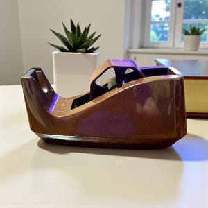 May include: A brown and tan tape dispenser with a curved design. The dispenser has a blade for cutting tape and a slot for the tape roll. A small succulent plant in a white pot is in the background.