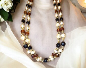 May include: A two-strand necklace featuring alternating beads in black, brown, white, and gold. The necklace is displayed on a white fabric, with a bouquet of white roses and a lit candle in the background.