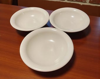 May include: Three white, shallow, round bowls are arranged on a dark wooden surface. The bowls have a simple, classic design with a slightly raised rim. The bowls are empty and appear clean, suggesting they are ready for use.